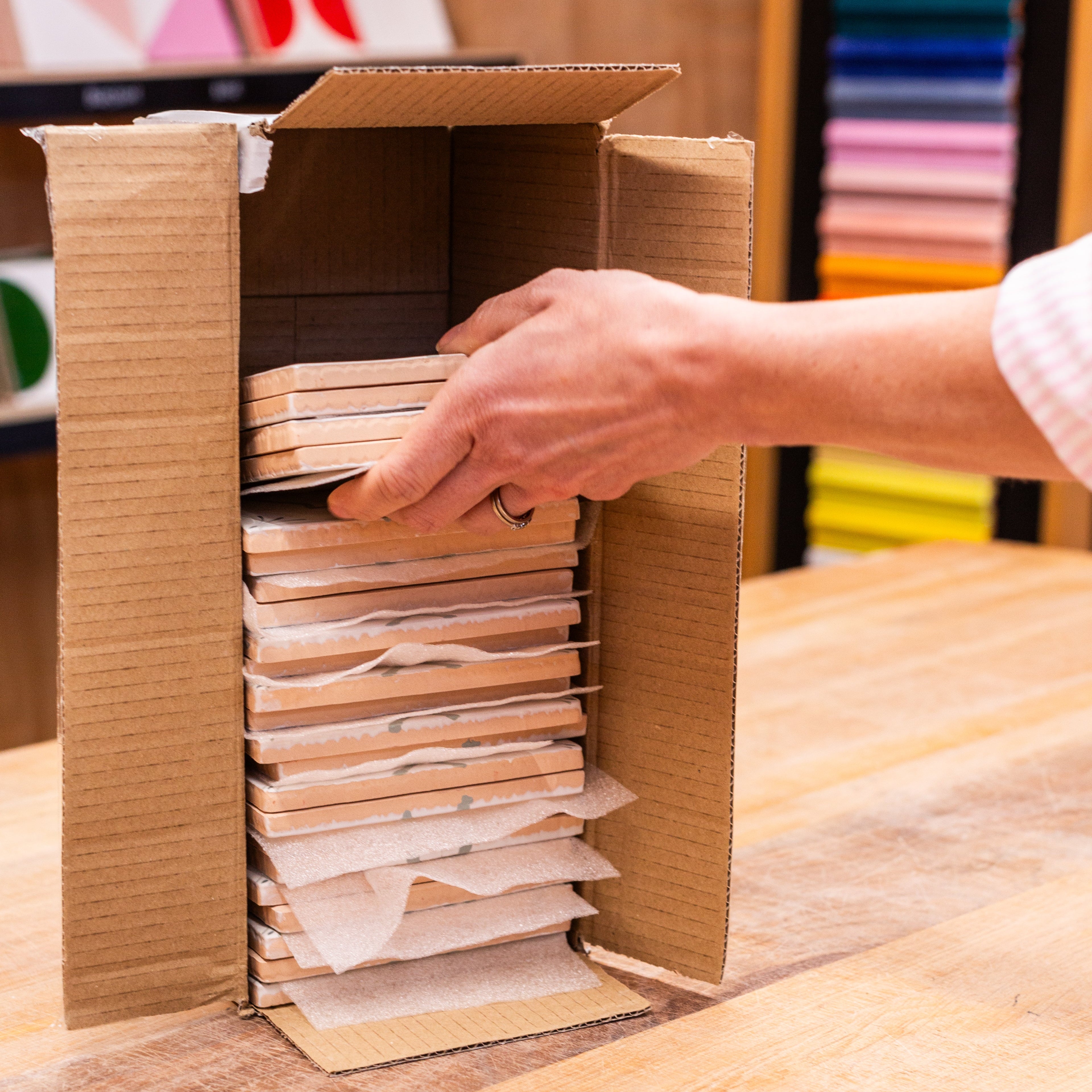 Person organizing a ceramic tile order in a cardboard box on a wooden table with color swatches in the background.