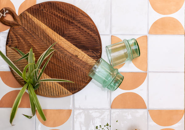 Bronze and white tiles with round chopping board, green glasses, wooden serving spoons and a leafy green plant