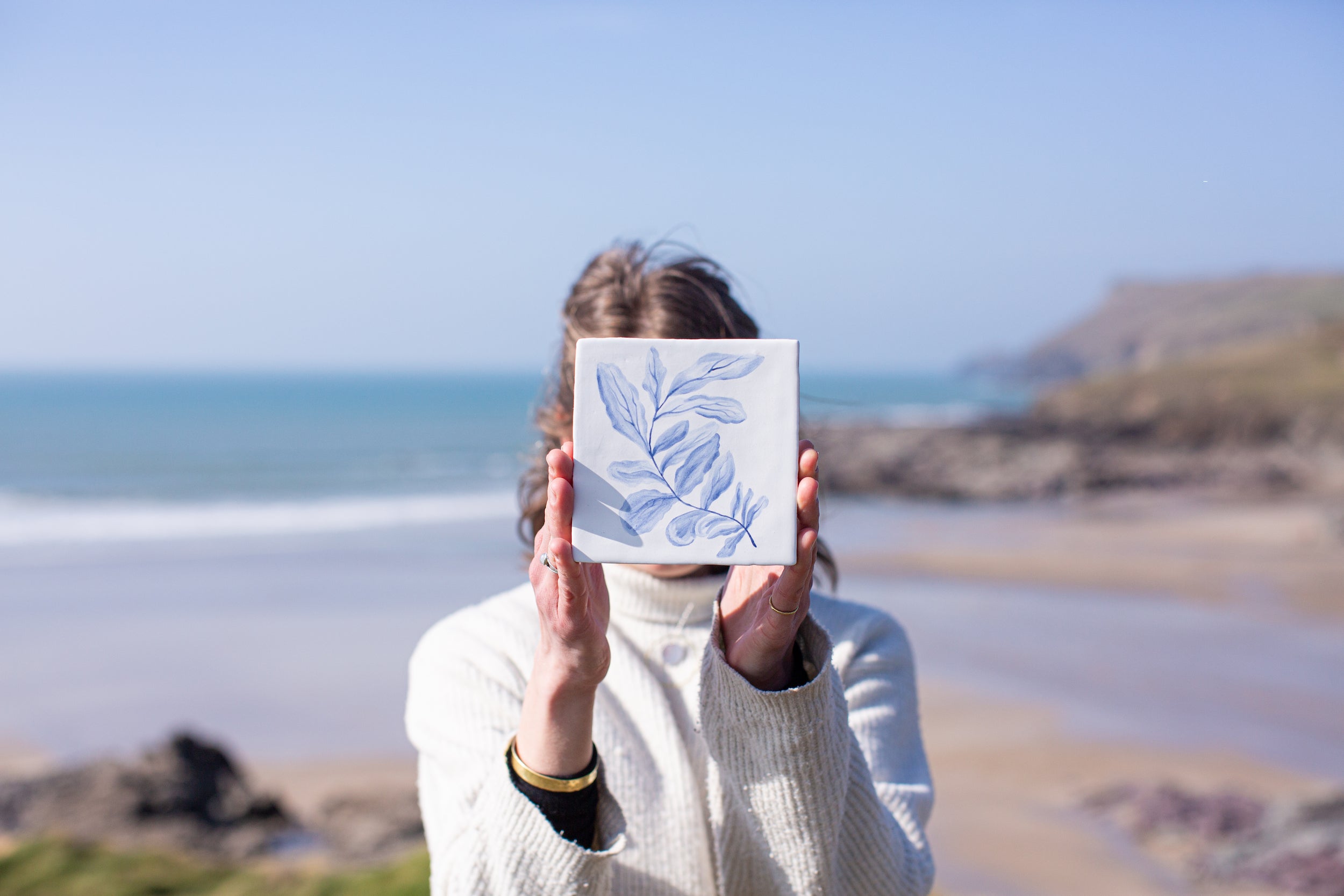 Person holding a ceramic tile with blue seaweed design in front of their face, with a Cornish beach in the background.