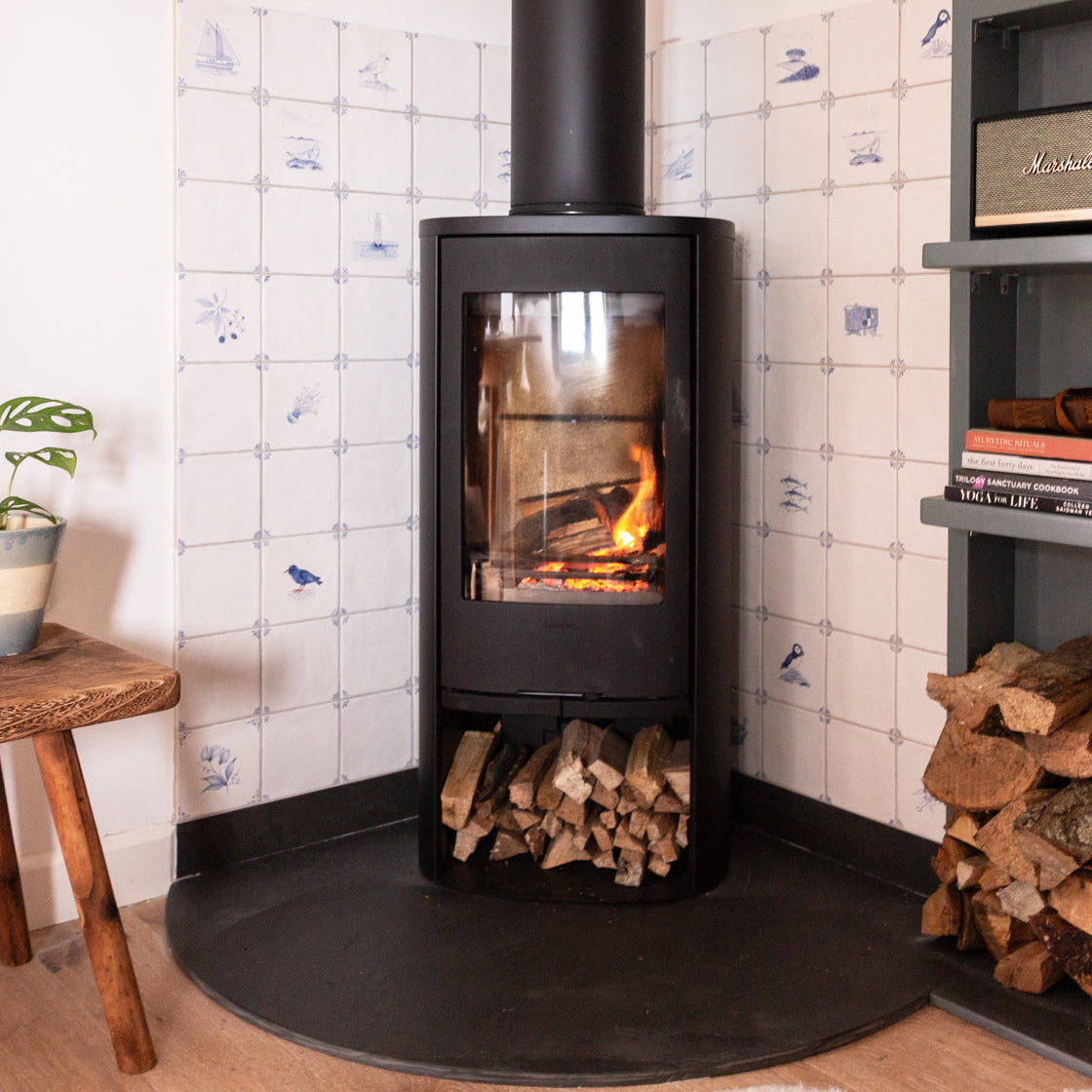 Wood-burning stove with fire lit, surrounded by stacked wood and a delft inspired tiled wall behind.